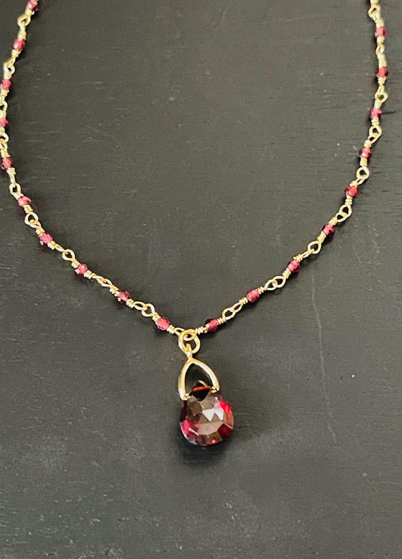 Puede incluir: Un delicado collar de cadena dorada con peque&ntilde;as cuentas rojas y un colgante de piedra roja.