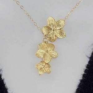 May include: A gold-colored necklace featuring three flower pendants of varying sizes. The flowers are detailed with textured petals and are suspended from a delicate chain. The necklace is displayed against a neutral background.