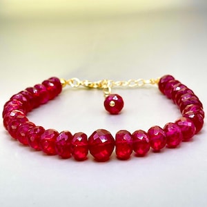 May include: A bracelet featuring a series of faceted, deep red beads arranged in a circular pattern. The bracelet includes a gold-toned metal chain with a lobster clasp and a single matching red bead dangling from the end of the extender chain.
