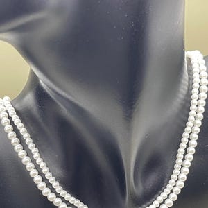 May include: A double-strand pearl necklace displayed on a black mannequin. The necklace features small, round, white pearls, creating a classic and elegant look. The strands are draped around the mannequin's neck, showcasing the jewelry's design.