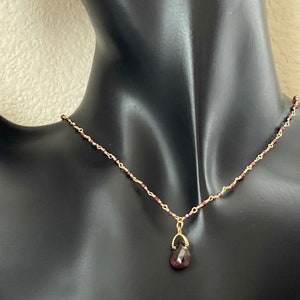 Puede incluir: Un collar de cadena dorada con un peque&ntilde;o colgante en forma de l&aacute;grima con una piedra preciosa de color rojo oscuro.