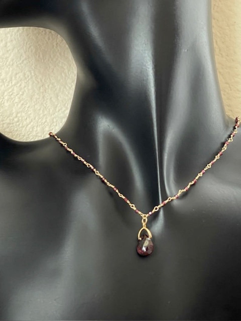 Puede incluir: Un collar de cadena dorada con un peque&ntilde;o colgante en forma de l&aacute;grima con una piedra preciosa de color rojo oscuro.