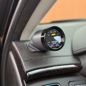 May include: A black and silver AEM air/fuel ratio gauge installed in a car's dashboard. The gauge displays numbers from 8 to 17, with the text "AIR/FUEL RATIO" and "MODE" visible. The dashboard is dark brown with a wood grain trim.