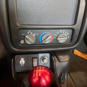 May include: Close-up of a red gear shift knob with the numbers 3, 5, R, 2, 4, and 6 printed on it. The knob is in a car's interior with a black dashboard and other controls.