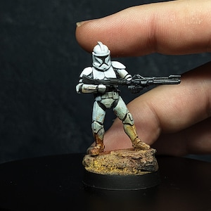 May include: A white and gray painted plastic action figure of a Star Wars clone trooper holding a black blaster rifle. The figure is standing on a small brown and yellow painted base.