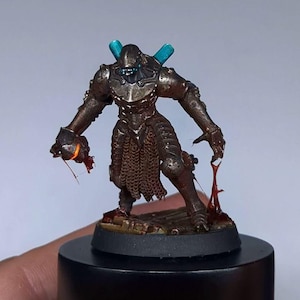 Könnte beinhalten: Miniaturfigur eines Kriegers in bronzefarbener Rüstung mit Kettenhemd und blauen Akzenten. Die Figur hält eine leuchtende Kugel in der einen Hand und Blut tropft aus der anderen. Der Sockel ist schwarz.