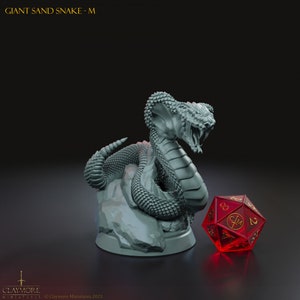 May include: A gray plastic miniature of a giant sand snake with its head raised and mouth open, posed on a rocky base. The snake is coiled around the base and has a textured, scaly surface. A red 20-sided die is in the foreground.