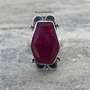 May include: A silver ring featuring a large, faceted, deep red gemstone. The gemstone is an elongated hexagon shape. The ring has decorative accents on the sides and the text "KusumJewellers" is visible at the top.