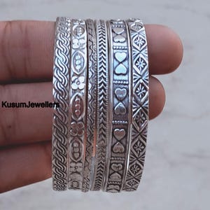 May include: A collection of silver bangles with intricate patterns, including floral, heart, and geometric designs. The bangles are stacked together, showcasing the variety of textures and styles. The brand name "KusumJewellers" is visible on the left side.