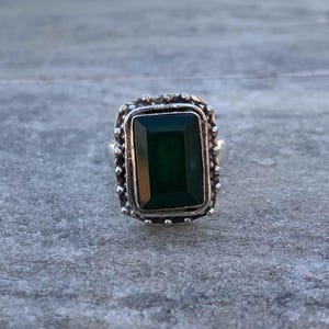 Puede incluir: Un anillo de plata con una gran gema rectangular de color verde esmeralda. La gema está engastada en un marco de plata decorativo y texturizado. El diseño del anillo es simple pero elegante, adecuado para el uso diario o ocasiones especiales.
