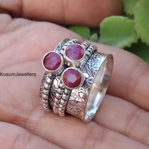 May include: A silver-toned ring with three round, red gemstones. The ring has a textured band with decorative patterns and additional spinning bands. The ring is shown on a hand.