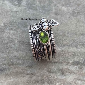May include: A silver ring with a bee motif and a green oval gemstone. The ring has detailed patterns and a spinning band. The text "Kusumjewellers" is visible on the ring.