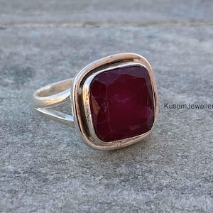 Puede incluir: Un anillo de plata con una piedra preciosa de color rojo oscuro de forma cuadrada engastada en un engaste de bisel. El anillo tiene una banda simple con un diseño dividido.
