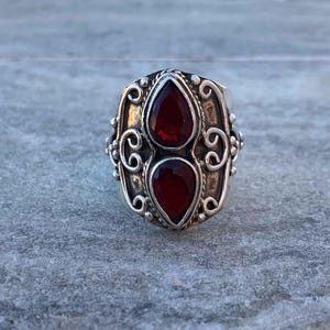 Könnte beinhalten: Ein silberner Ring mit zwei tropfenförmigen, tiefroten Edelsteinen. Der Ring hat ein verziertes Design mit Wirbelmustern und kleinen Perlenakzenten um die Edelsteine. Der Ring ist vor einem neutralen Hintergrund platziert.