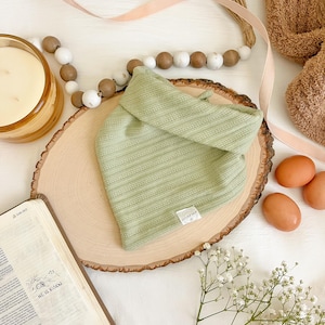 May include: A green ribbed fabric baby bandana with a small white tag that says "UNAFRAID" on a wooden slice. The bandana is surrounded by white and brown wooden beads, a brown towel, a candle, a book with the words "HE IS RISEN" on the cover, and white flowers.