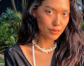 May include: A woman with long dark hair wearing a black sheer top with a white tie and a white shell necklace.