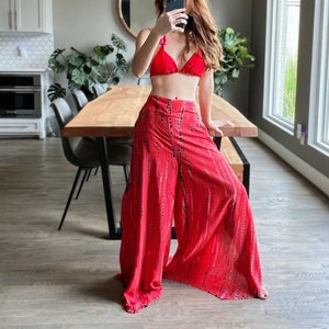 以下が含まれることがあります： 赤いビキニトップと黒と白のタイダイ柄の赤いワイドレッグパンツを着用した女性。彼女は木製のテーブルと椅子のあるキッチンに立っています。