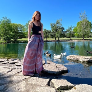 May include: A person wearing a black one-piece swimsuit and pink and purple tie-dye pants stands on a rocky path by a pond.  Ducks are swimming in the pond.