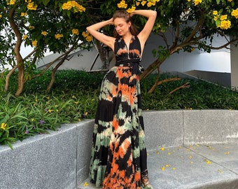Op de afbeelding: Een vrouw draagt een zwarte en oranje tie-dye maxi-jurk met een halternek en een zwarte riem. Ze staat voor een gele bloeiende struik.