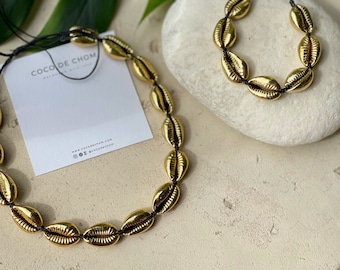 May include: Two gold shell necklaces with black string. The larger necklace is on a white card with the text "Coco de Chom" and website information. The smaller necklace is on a light gray stone.