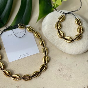 May include: Two gold shell necklaces with black string. The larger necklace is on a white card with the text "Coco de Chom" and website information. The smaller necklace is on a light gray stone.