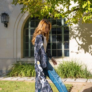 May include: A woman wearing a long, open-front kimono with a dark blue background and a white crane pattern. The kimono is paired with blue jeans and white heels. The background includes a building with a large arched window and green foliage.