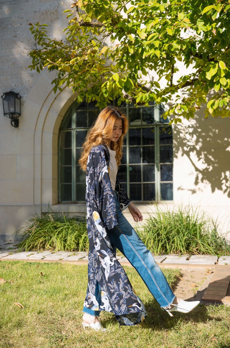 May include: A woman wearing a long, open-front kimono with a dark blue background and a white crane pattern. The kimono is paired with blue jeans and white heels. The background includes a building with a large arched window and green foliage.