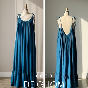 Back view of a blue turquoise open back maxi dress with tie shoulders and a flowing oversized fit, worn barefoot on the beach.