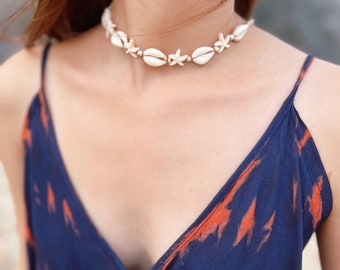 May include: A white seashell necklace with starfish accents. The necklace is worn by a person with a blue and orange tie-dye top.