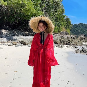 Op de afbeelding: Een vrouw in een rode maxi-jurk met een strik om de hals en een brede strohoed loopt op een wit zandstrand.
