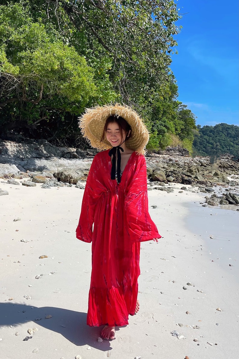 Op de afbeelding: Een vrouw in een rode maxi-jurk met een strik om de hals en een brede strohoed loopt op een wit zandstrand.