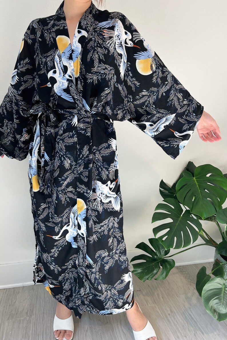 May include: Black kimono-style robe with a crane and foliage pattern. The robe features white, blue, and yellow cranes set against a backdrop of gray leaves and yellow suns. It has wide sleeves and a matching tie belt.