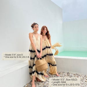 Two women wearing long, flowy, white and black tie-dye dresses. The women are standing on a gravel surface in front of a white wall and a small pool of water. One woman is holding a woven straw bag.