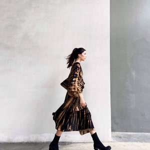 May include: A woman wearing a brown and black tie-dye dress with long sleeves and a flowing skirt. She is walking towards the right side of the image, wearing black ankle boots with chunky heels.