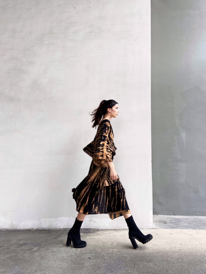 May include: A woman wearing a brown and black tie-dye dress with long sleeves and a flowing skirt. She is walking towards the right side of the image, wearing black ankle boots with chunky heels.
