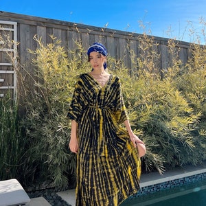 May include: A woman wearing a yellow and black tie-dye maxi dress with a blue headscarf. She is standing by a pool with a wooden fence and bamboo plants in the background.
