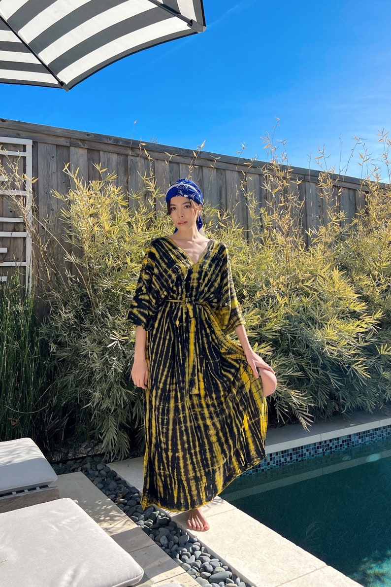 May include: A woman wearing a yellow and black tie-dye maxi dress with a blue headscarf. She is standing by a pool with a wooden fence and bamboo plants in the background.