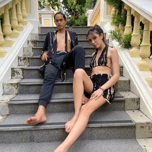 May include: A couple poses on a set of stone stairs. The woman is wearing a black and white tie-dye halter top and matching shorts. The man is wearing a black and white tie-dye robe over black pants.
