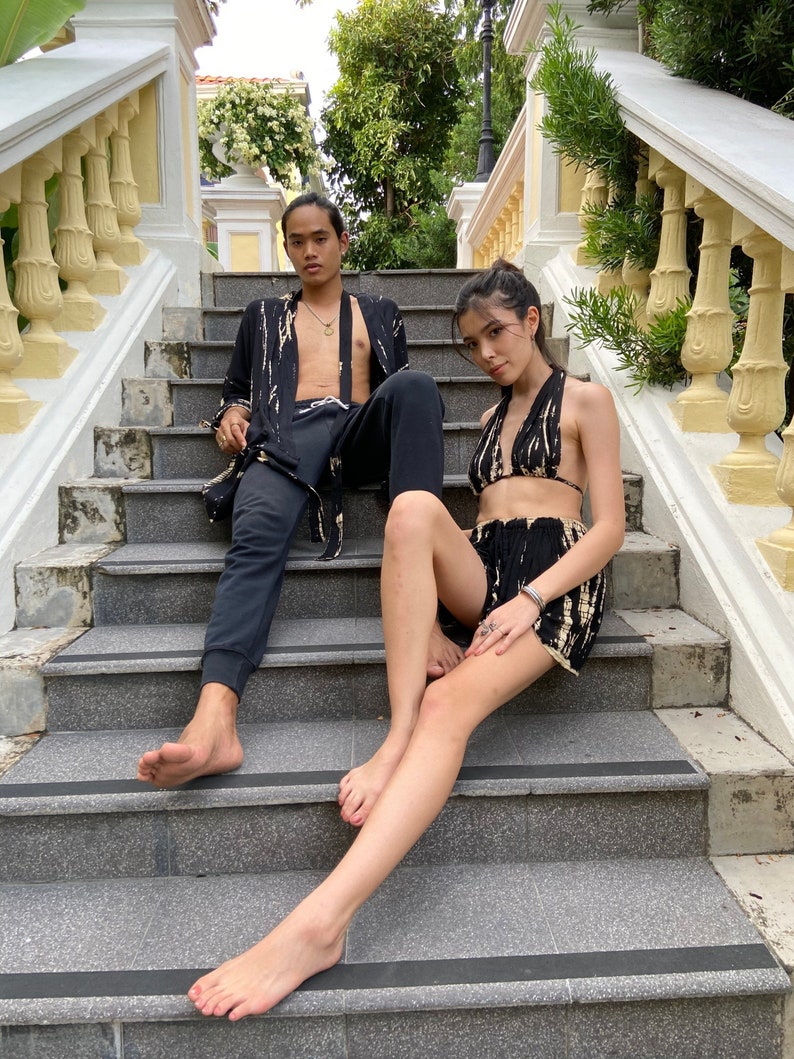 May include: A couple poses on a set of stone stairs. The woman is wearing a black and white tie-dye halter top and matching shorts. The man is wearing a black and white tie-dye robe over black pants.