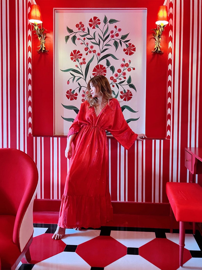 Op de afbeelding: Een vrouw in een rode maxi-jurk staat in een kamer met rood-wit gestreepte muren. De muren zijn beschilderd met een bloemenmotief. De vrouw kijkt naar links.