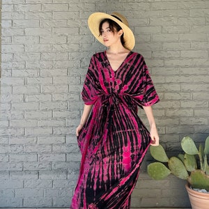 May include: A woman wearing a pink and black tie-dye maxi dress with a straw hat. The dress has a V-neck and a tie at the waist.