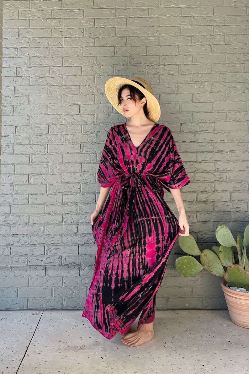 May include: A woman wearing a pink and black tie-dye maxi dress with a straw hat. The dress has a V-neck and a tie at the waist.