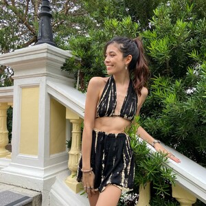 May include: A woman wearing a black and white tie-dye halter top and matching shorts. The outfit is a two-piece set with a cropped top and high-waisted shorts. The woman is standing on a set of stairs with a lush green bush behind her.