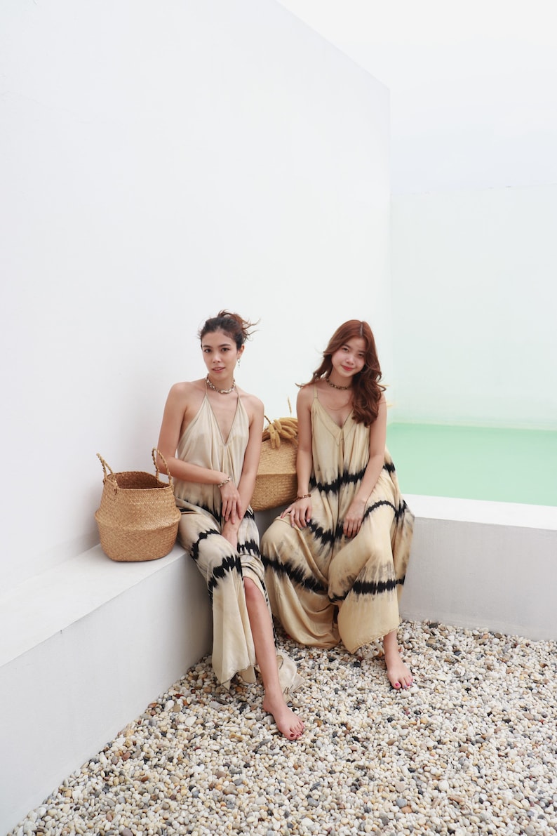 May include: Two women wearing long, flowy, tie-dye dresses with black stripes. They are sitting on a white ledge with a woven basket beside them. The background is a white wall and a light blue surface.