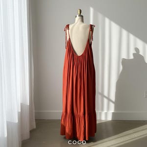 model in burn orange open back maxi dress with a low back and tie straps. The maxi dress has a tiered skirt, resort wear.