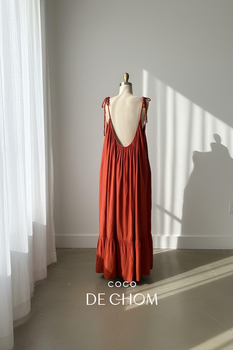 model in burn orange open back maxi dress with a low back and tie straps. The maxi dress has a tiered skirt, resort wear.