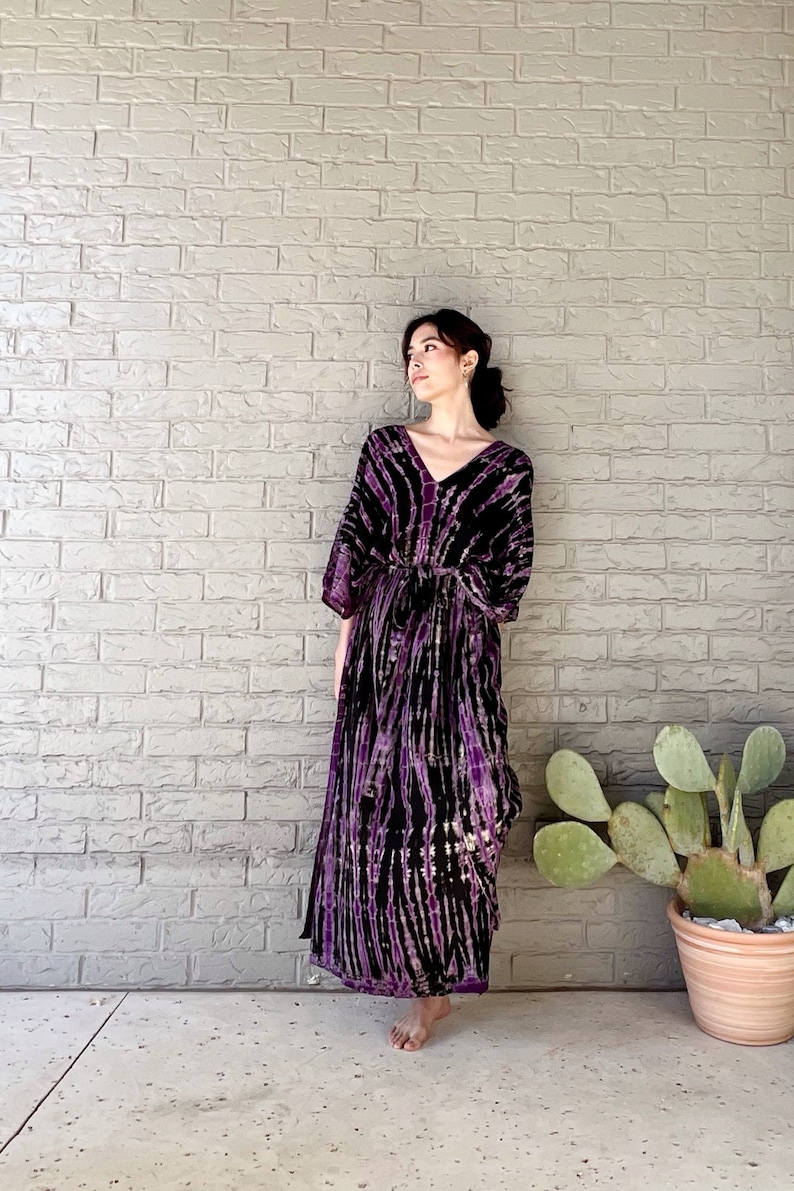 May include: A long, purple and black tie-dye maxi dress with a V-neckline and 3/4 sleeves. The dress has a tie at the waist and is made of a soft, flowing fabric.