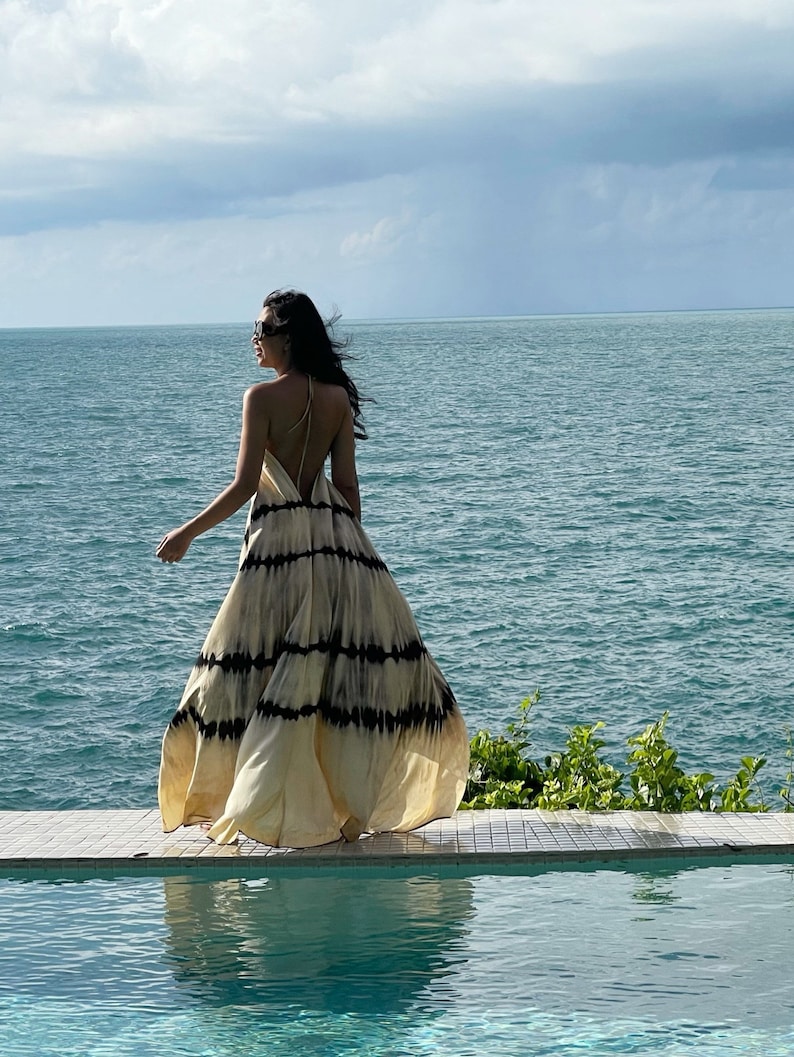 May include: A woman in a white and black tie-dye maxi dress walks towards the ocean. She is wearing sunglasses and has her back to the camera. The ocean is a deep blue and the sky is cloudy.