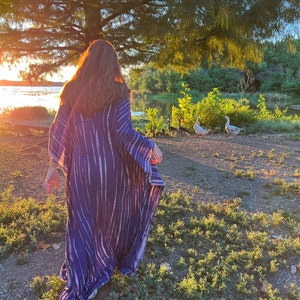 May include: A long, flowing blue and white tie-dye patterned dress. The dress is worn outdoors near a body of water, with a tree and geese in the background. The sun is setting, casting a warm glow over the scene.