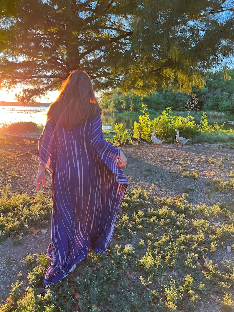 May include: A long, flowing blue and white tie-dye patterned dress. The dress is worn outdoors near a body of water, with a tree and geese in the background. The sun is setting, casting a warm glow over the scene.